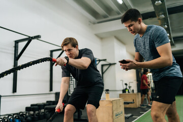 Portrait of dedicated sportsman exercising with battle rope while coach encouraging during cross training in health club standing with stopwatch. Concept of healthy lifestyle, physical sport activity.