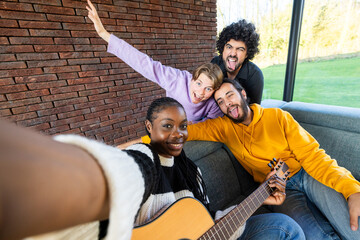 Capturing a moment of pure joy, this image shows a group of diverse friends taking a selfie. One plays a guitar, while the others show playful expressions. They are seated against a brick wall inside - obrazy, fototapety, plakaty