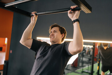 Tired motivated beginner sportsman showing hard expression on face in pulling up own body. Exhausted muscular male hanging on bar and does one pull-up during sport workout training in gym.