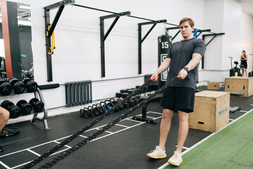 Motivated beginner sportsman doing battle ropes functional training, pulling ropes, exercising strength at modern gym, looking at camera. Strong male athlete working out with battling rope at sport