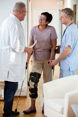 An elderly female patient talking with doctors in clinic hall. Woman wearing bondage, medical splint, knee brace on the leg, communicating with the attending physician at a hospital. Signing insurance