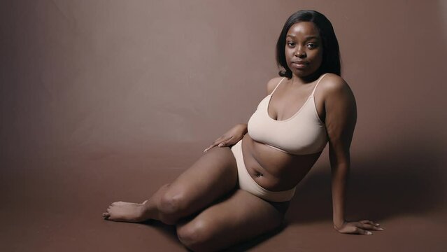 Studio shot of young pretty African American woman with curvy body wearing minimalist lingerie sitting on floor and looking at camera while posing on coffee color cyc wall