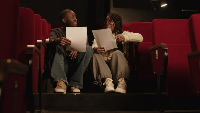 Low angle shot of young Black couple of actors or standup comedians sitting on staircases in theatre with red seats and chatting while revising monologue before stage performance