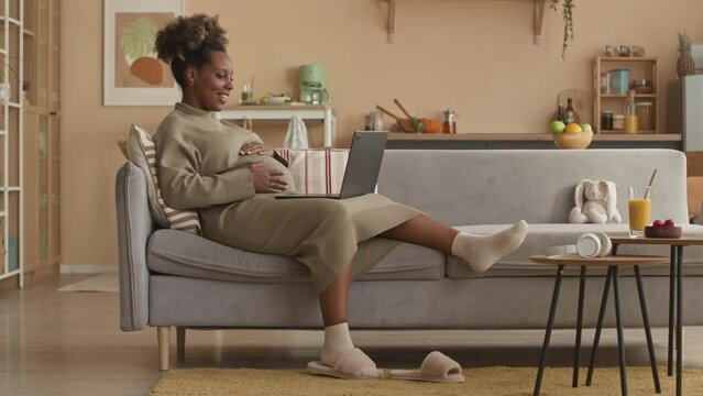 Young happy Black woman expecting baby sitting in relaxed pose on couch in bright modern apartment having video chat on laptop