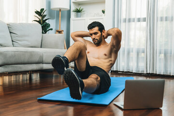 Athletic and sporty man doing crunch on fitness mat while follow online home workout exercise...