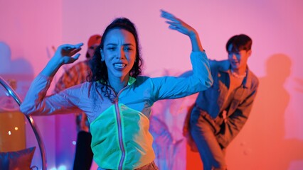 Attractive happy dancer looking at camera while smart woman moving at rhyme with neon light. Professional hispanic performer break dancing while wearing colorful cloth with diverse friend. Regalement.