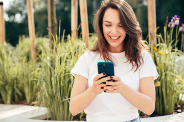 Young beautiful smiling hipster woman in trendy summer casual clothes. Carefree woman posing in the street. Positive model holds mobile phone, looks at cellphone screen, uses smartphone apps