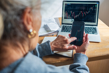elderly retired woman senior trading on line with smartphone and laptop investment bitcoin stock...