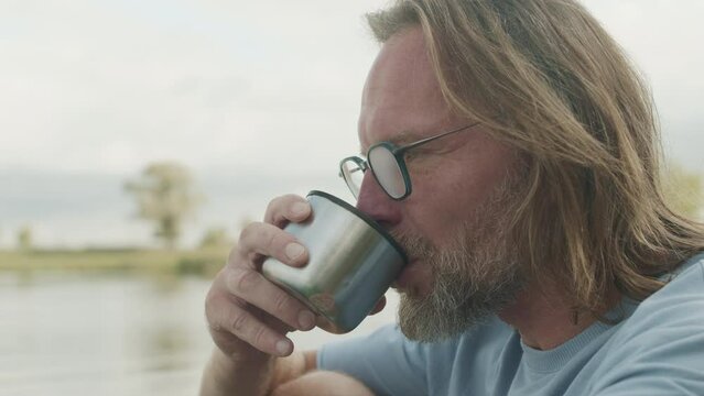 Chest up of Caucasian mid-aged man drinking tea from thermos cup while sitting by water, enjoying nature and landscape