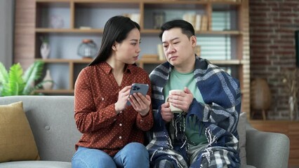 Asian family couple uses online medical care application using mobile phone sitting on sofa in living room at home. Wife supports a sick husband by searching for a remote consultation with a doctor