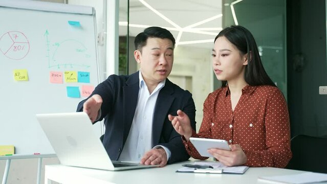 Asian boss consult young female worker at workplace in business office. Male manager advises, talking to an employee about a development project. Executive explains work issues using laptop computer