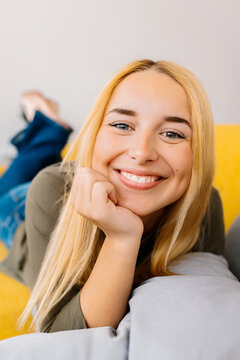 Vertical portrait of lovely young blonde woman looking at camera relaxing on sofa at home