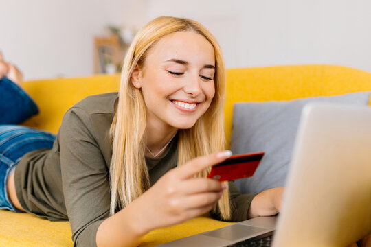 Happy cheerful smiling young blonde woman doing online shopping using credit card and laptop computer. Millennial girl making online payment.