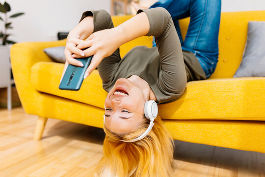 Happy young girl watching social media content at smart phone device. Millennial blonde woman having fun using cellphone relaxing on sofa at home