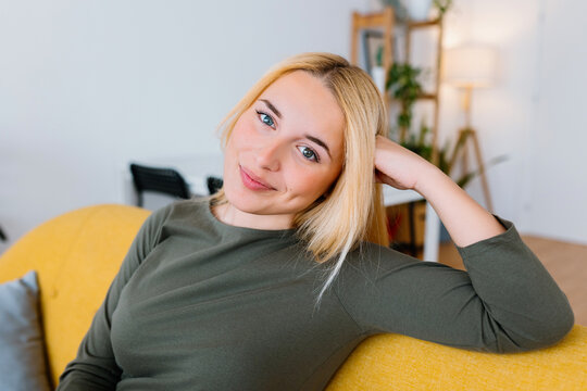 Happy young woman sitting on sofa at home. Smiling portrait of beautiful blonde girl looking at camera