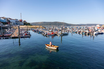 A serene view of a busy harbor in Fisterra Spain filled with various boats, flanked by a town,...