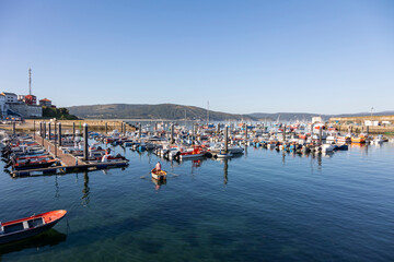 A serene view of a busy harbor in Fisterra Spain filled with various boats, flanked by a town, under the vast expanse of a clear blue sky, depicting tranquility and daily life - obrazy, fototapety, plakaty