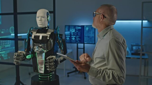 Medium shot of mid adult Caucasian male engineer using digital tablet while activating humanoid robot in laboratory