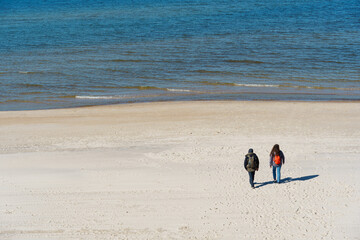 A couple of travelers in warm clothes with backpacks walk along a deserted beach near the sea in...