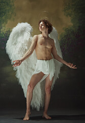 Ethereal Charm. Young man with curly hair and white wings poses with air of grace against vintage background. Concept of fashion and beauty, modern art and historical fiction fusion. Ad
