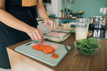 A woman is preparing food on a wooden table, she is cutting tomatoes and ham on a cutting board