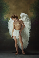 Young man with curly hair and celestial wings posing shows earthly beauty against vintage studio background. Concept of fashion, modern art and historical fiction fusion. Ad