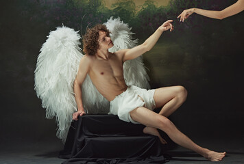 Renaissance painting come to life. Man with curly hair sits with large wings spread out behind him, reaching out to hand against vintage studio background. Concept of modern art and historical fiction