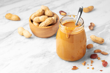 Natural peanut butter in glass jar with spoon and peanuts in wooden bowl on white marble table