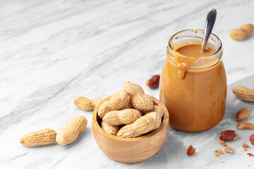 Peanuts in wooden bowl and peanut butter in glass jar on marble table