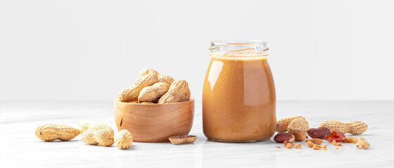 Peanut butter in jar and roasted peanuts in wooden bowl on white marble table