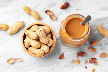 Peanut butter in glass jar with spoon and peanuts in wooden bowl on white marble table