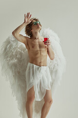 Man with wings and curls in modern sunglasses throws his head back in moment of carefree joy holds drink in hand against white studio background. Concept of modern art and historical fiction fusion.