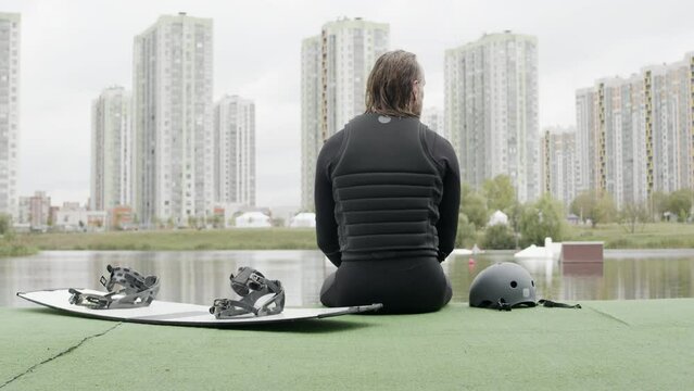 Zoom-out back view medium shot of long-haired male wakeboarder in wetsuit sitting by water on green dock with board and helmet besideZoom-out back view medium shot of long-haired male wakeboarder in w