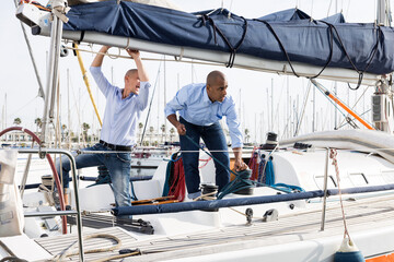 Two men in blue shirts and jeans with bare legs drinking beer and having fun on a yacht in seaport - 795119929