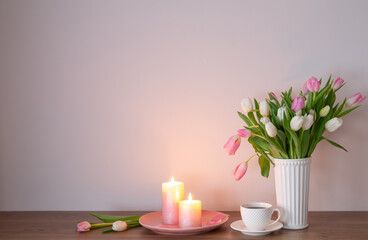 cup of coffee and spring tulips in vase with burning candles on wooden shelf