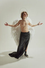 Man with curly hair, white wings and halo above his head posing with arms open wide against white studio background. Concept of fashion and beauty, modern art and historical fiction fusion. Ad