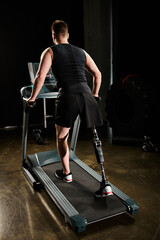 A man with a prosthetic leg is walking on a treadmill in a dimly lit room, focusing on his workout...