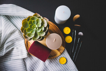 Candles, wooden board, makeup brushes, textile and cactus on a black background. Copy space
