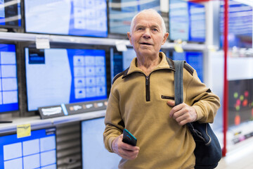 Elderly grayhaired man pensioner looking counter with modern digital televisors in showroom of digital goods store - 795125153