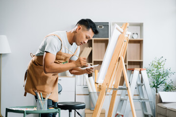Asian male painter do artwork in art workshop, painting supplies, oil pastels, two canvas easel, creative space with paintbrush in art studio