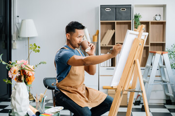 Asian male painter do artwork in art workshop, painting supplies, oil pastels, two canvas easel, creative space with paintbrush in art studio