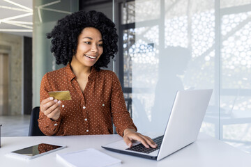 Cheerful young woman with curly hair using a credit card for an online purchase. Sitting at a desk...