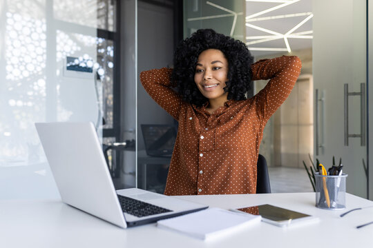 A young African American woman in a polka dot shirt stretches comfortably at her modern office desk, exuding a relaxed, confident vibe.