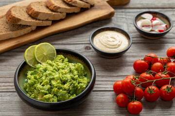 Fresh chunky guacamole decorated with slices of lime,  homemade bread slices on a wooden kitchen board, cherry tomatoes, tahini sauce, and red radishes on the table. Homemade, healthy vegan meal. 