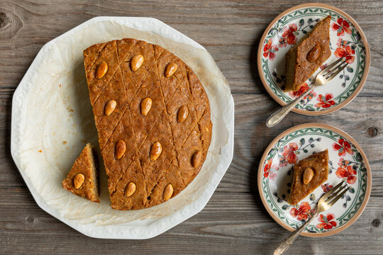 Top view of round cake with almonds, fenugreek seeds, and semolina and two plates with pieces of this cake. Sweet traditional helbeh (fenugreek cake) dessert. 