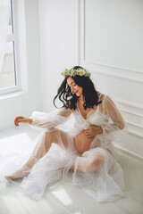Attractive pregnant woman in white polka dot peignoir and gypsophila wreath sitting on the floor...