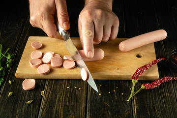 The cook cuts the sausage into small pieces with a knife. Preparing a delicious breakfast with milk sausage and spices on the kitchen table in a tavern - obrazy, fototapety, plakaty