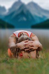 Young active barefoot woman bending forward while stretching on green grass on mountain landscape, Yoga amid peaks: Young woman’s body workout thrives in the mountain summer retreat