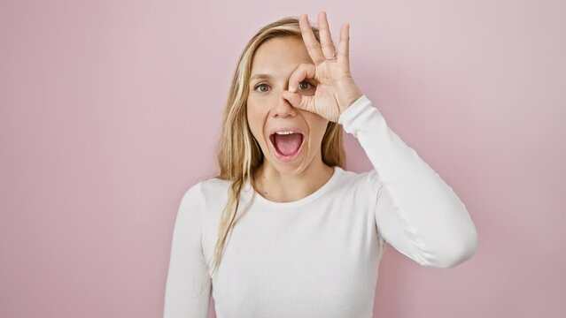 Blonde bombshell radiates shock and surprise! gutsy young woman makes a cheeky 'ok' gesture, peeks through fingers over an isolated pink wall background.