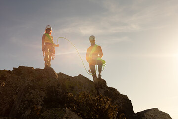Two people are standing on a mountain, one of them holding a rope. The scene is bright and sunny,...